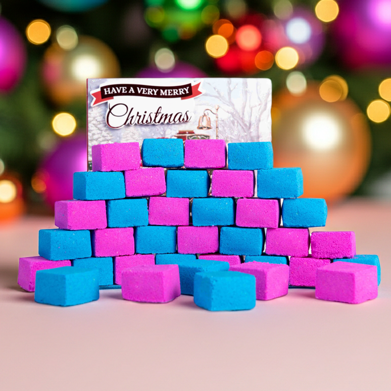 Stack of pink and blue bath  bomb blocks with a Christmas-themed card in front of a decorated Christmas tree.