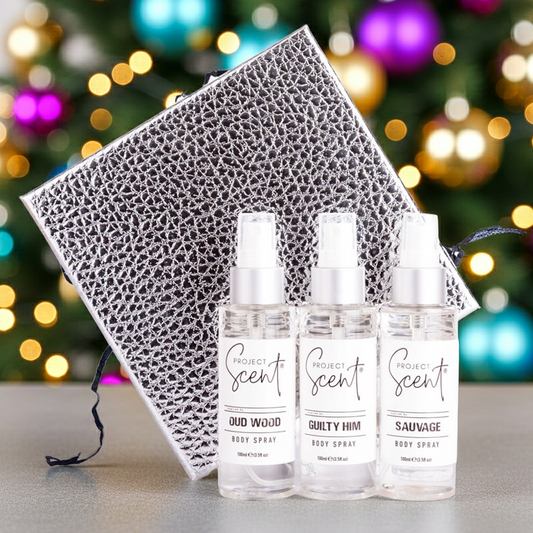 Three body spray bottles with a decorative box in front of a Christmas tree.