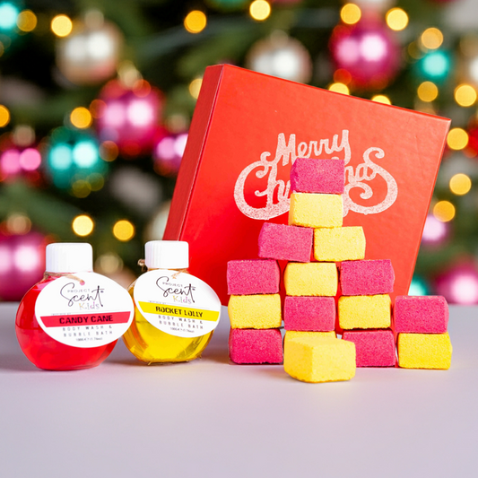 Colorful bath bomb blocks, a red gift box with 'Merry Christmas' text, and small bottles of body wash on a blurred Christmas tree background.