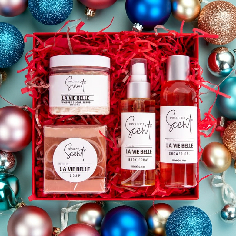 Flatlay of Christmas body care gift set surrounded by festive decorations. Features handmade sugar scrub, body spray, soap bar and shower gel gift box.