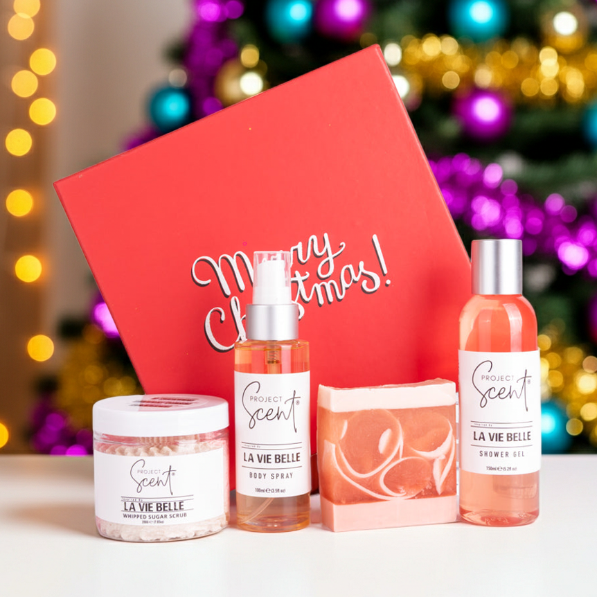 Christmas body care gift set displayed in front of a Christmas tree. Includes handmade sugar scrub, body spray, soap bar and shower gel in festive packaging.