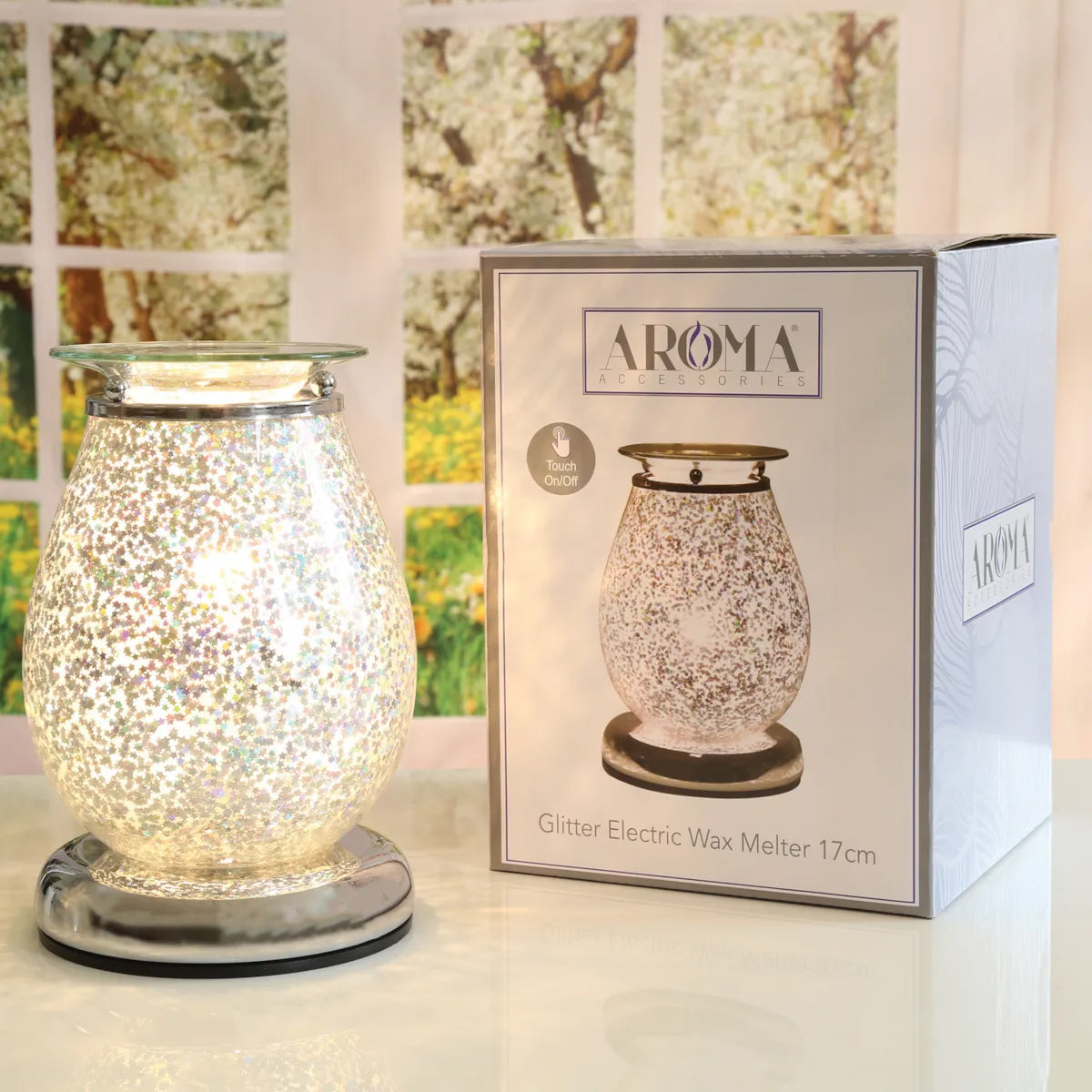 A glittery electric wax warmer with a clear glass body and iridescent silver sparkle finish, shown next to its product packaging box on a white surface.