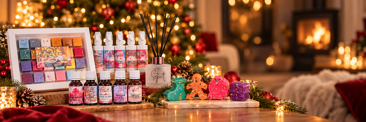 A festive holiday gift set displayed on a wooden table in a cozy living room setting with a burning fireplace and Christmas tree in the background. The set includes several small bottles with colorful labels, a framed multicolor card or box, a reed diffuser with reeds, and several bath bombs in green, gingerbread man, pink, and purple.
