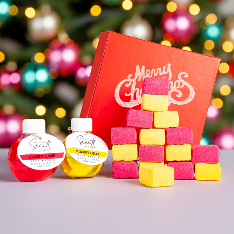 Colorful bath bomb blocks, a red gift box with 'Merry Christmas' text, and small bottles of body wash on a blurred Christmas tree background.
