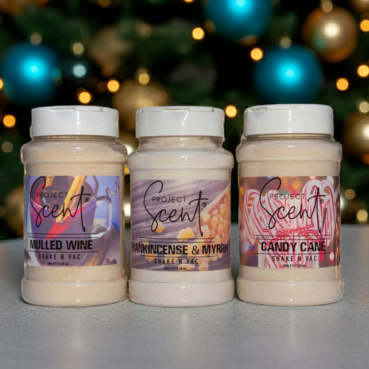 Christmas Shake N Vac carpet freshener displayed in front of a Christmas tree. Available in festive scents such as Gingerbread, Christmas Tree and Holly Jolly.